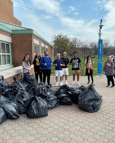 Campus Cleanup Students standing in front of 1P after finishing the Campus Clean Up. There are 20+ black garbage bags in front of them on the floor. 